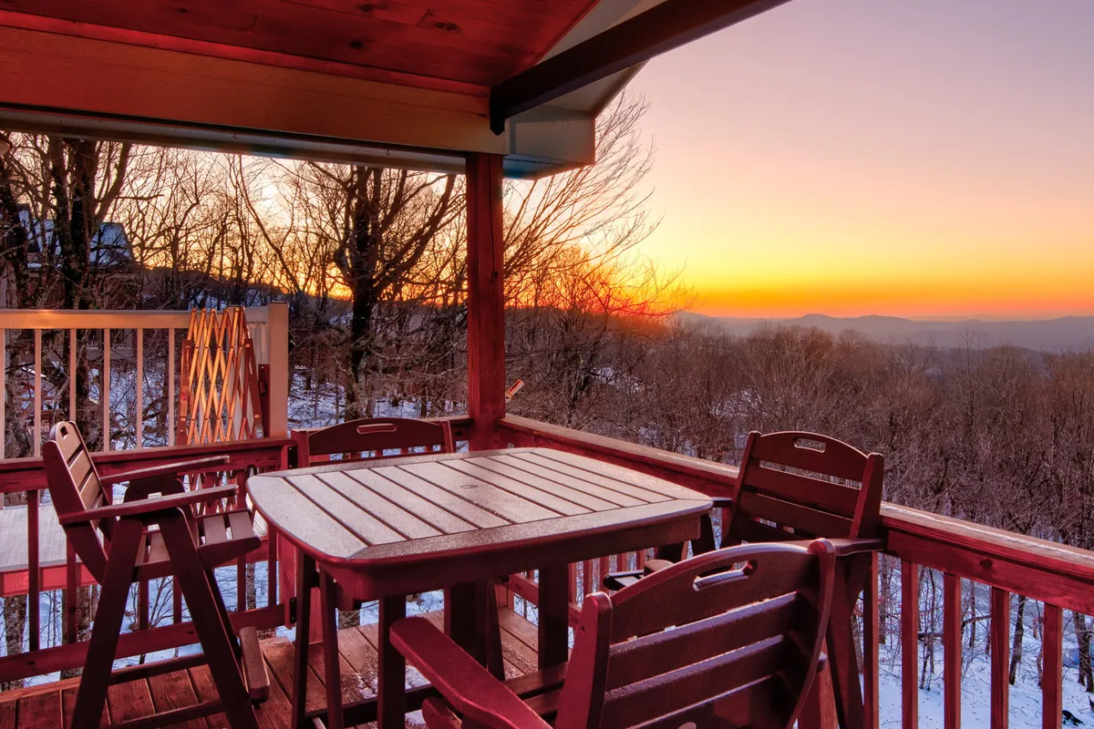 The image captures an inviting outdoor dining area with a wooden table and chairs set against the backdrop of a breathtaking mountain sunset. Silhouetted trees frame the expansive view, suggesting tranquility and the perfect spot for enjoying meals or relaxing with loved ones as day turns to night.