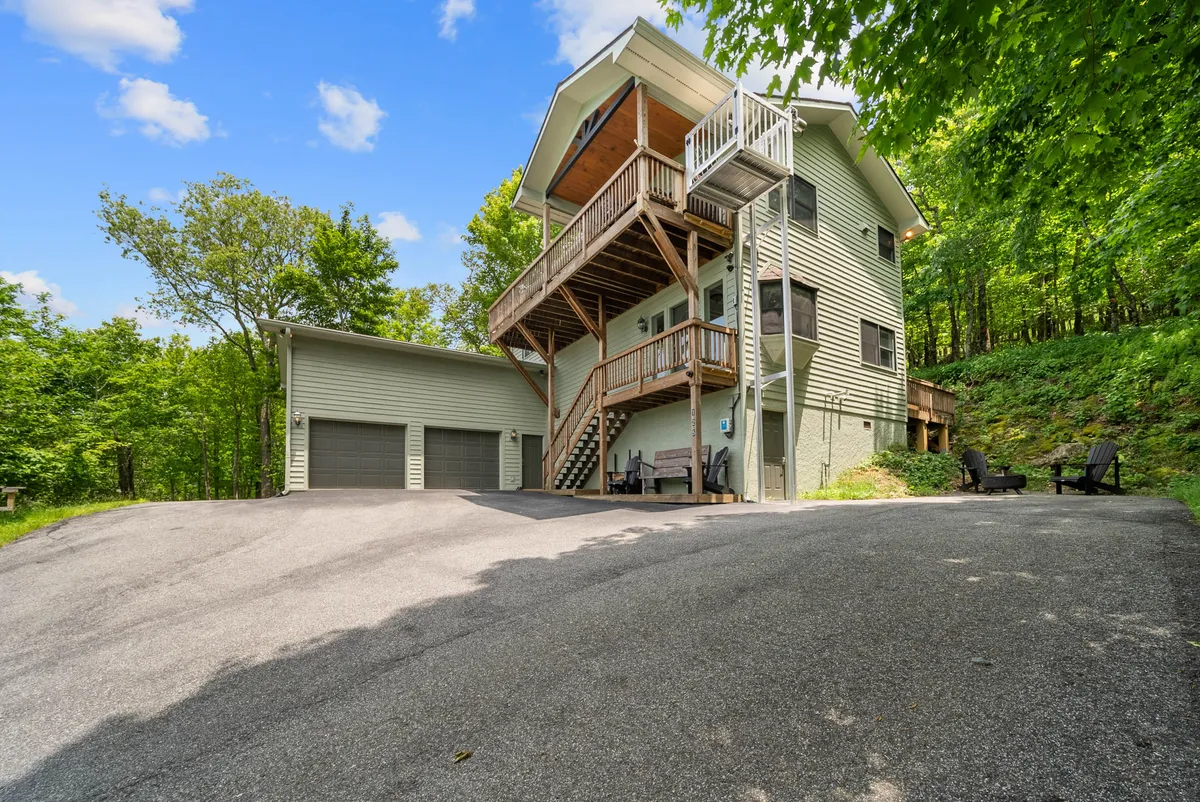 A welcoming view of the home's multi-level exterior, featuring a spacious driveway, a wooden balcony, and lush greenery. The image captures the essence of arriving at the property and highlights the inviting outdoor spaces ideal for gatherings and nature appreciation throughout the year.