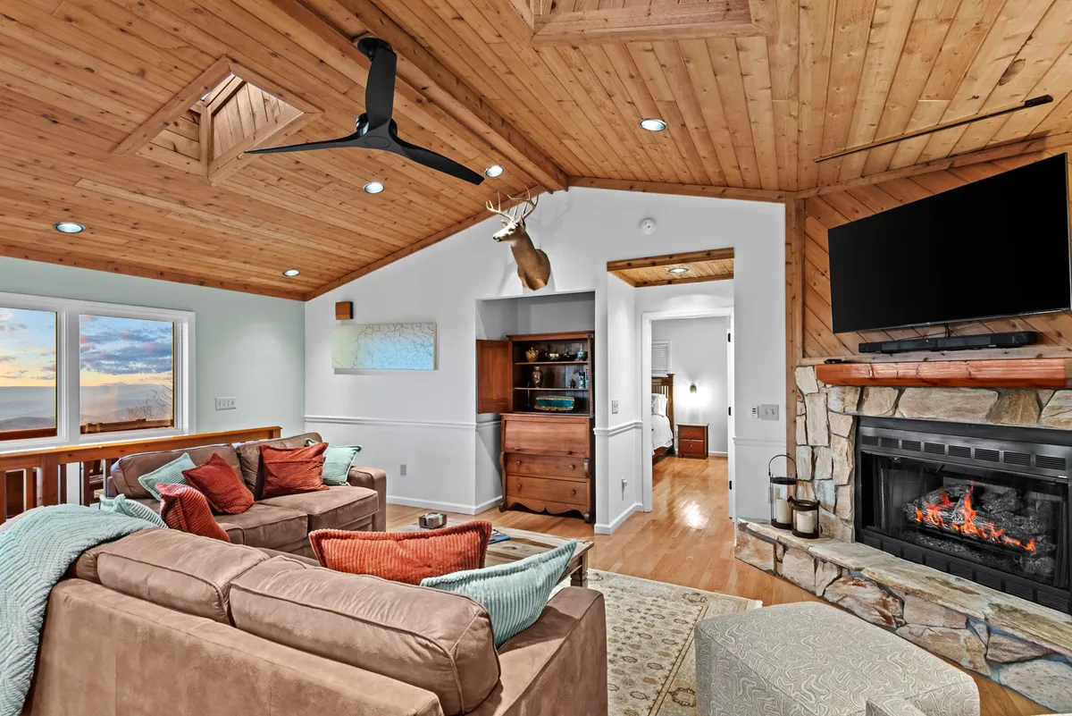 A sun-filled living room with a comfortable sofa adorned with colorful cushions, a warm fireplace, rich wooden ceiling, and large windows letting in abundant natural light. The space is inviting for both work and relaxation, with an atmosphere suitable for group brainstorming or quiet focus.