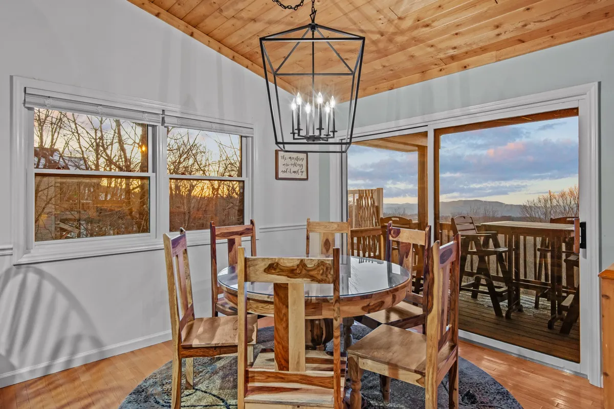 A cozy dining area highlighted by a rustic chandelier, a spacious table, and large windows offering a sunset view. The sliding door leads directly to the deck, inviting guests to enjoy fresh mountain air and nature breaks.