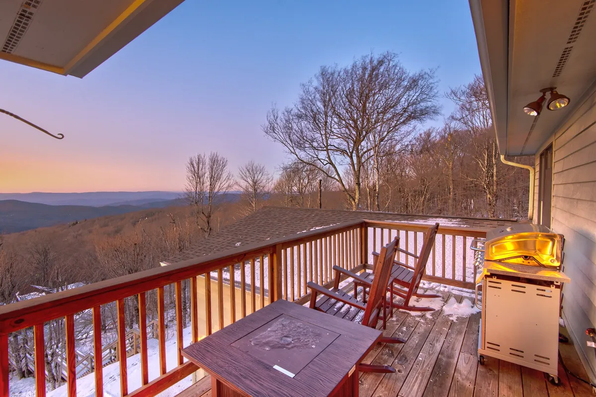This image captures a spacious wooden deck outfitted with comfortable chairs and a BBQ grill, set against a sweeping mountain view. The deck is elevated, providing a perfect perch to enjoy the scenery, relax, or have a meal outdoors. The warm light and tranquil setting evoke a peaceful, contemplative mood ideal for both gatherings and quiet moments.