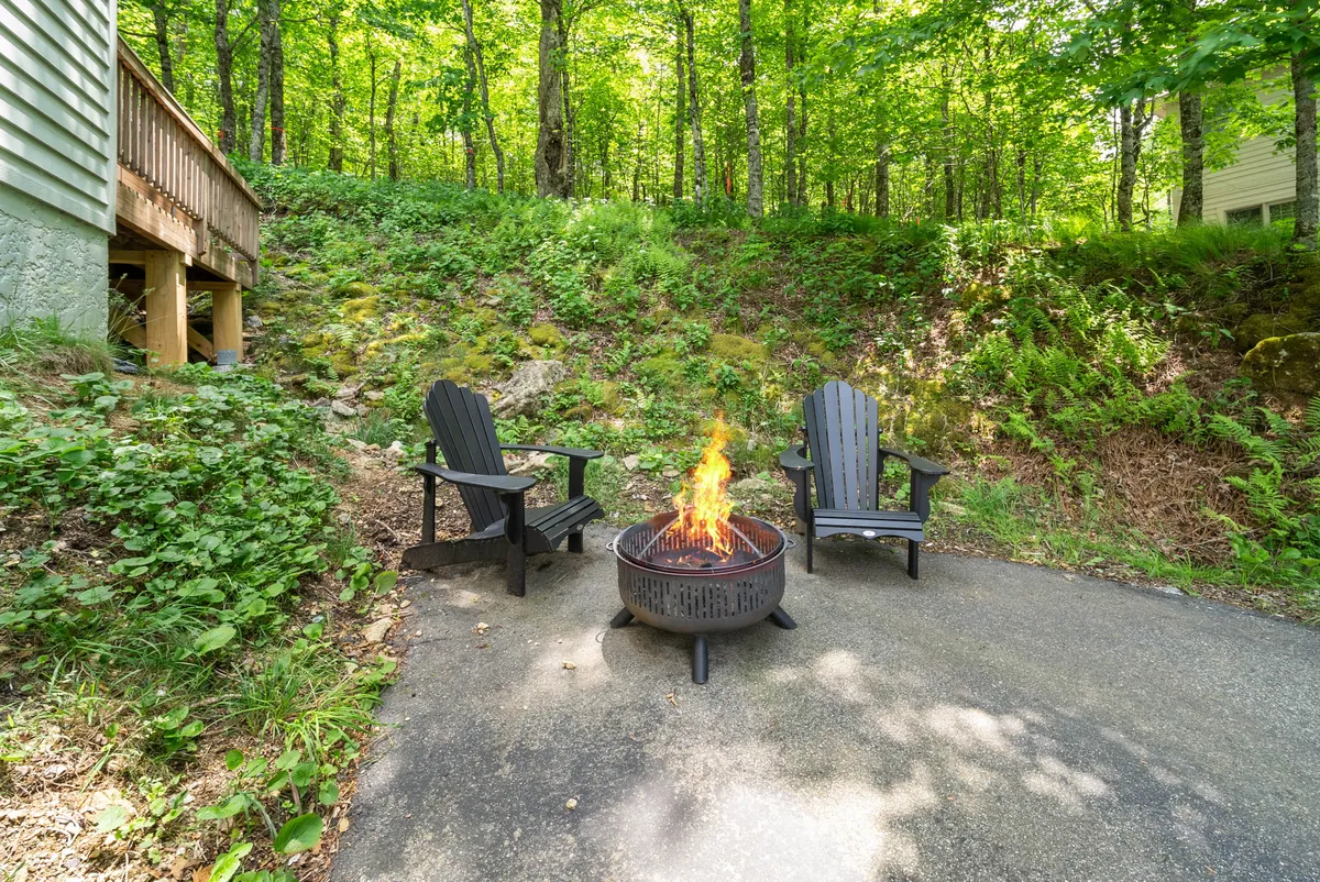 This cozy exterior setting features a fire pit surrounded by Adirondack chairs, with greenery all around. The scene is ideal for evening storytelling, stargazing, or gathering with friends and family, creating a warm, inviting outdoor atmosphere.