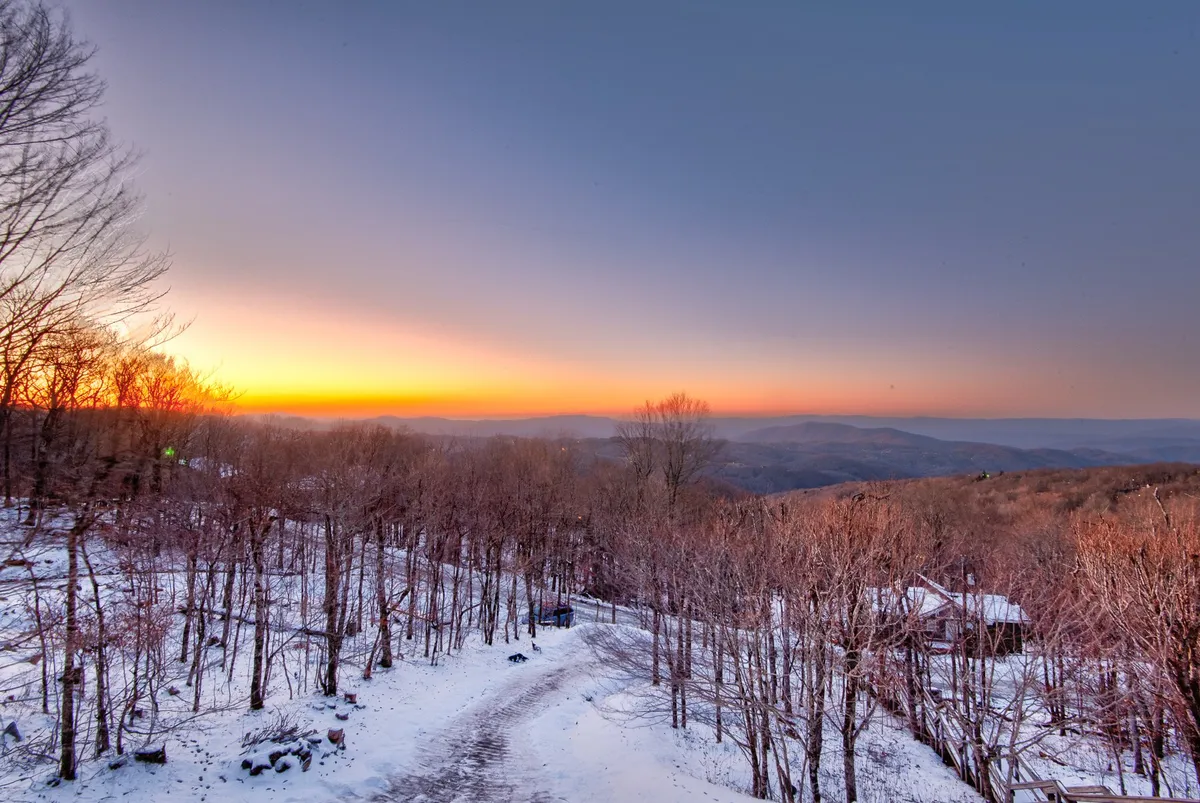 A panoramic mountain landscape bathed in sunset light, with snow-covered ground and silhouetted trees. The peaceful scene evokes tranquility and an immersive connection to nature.