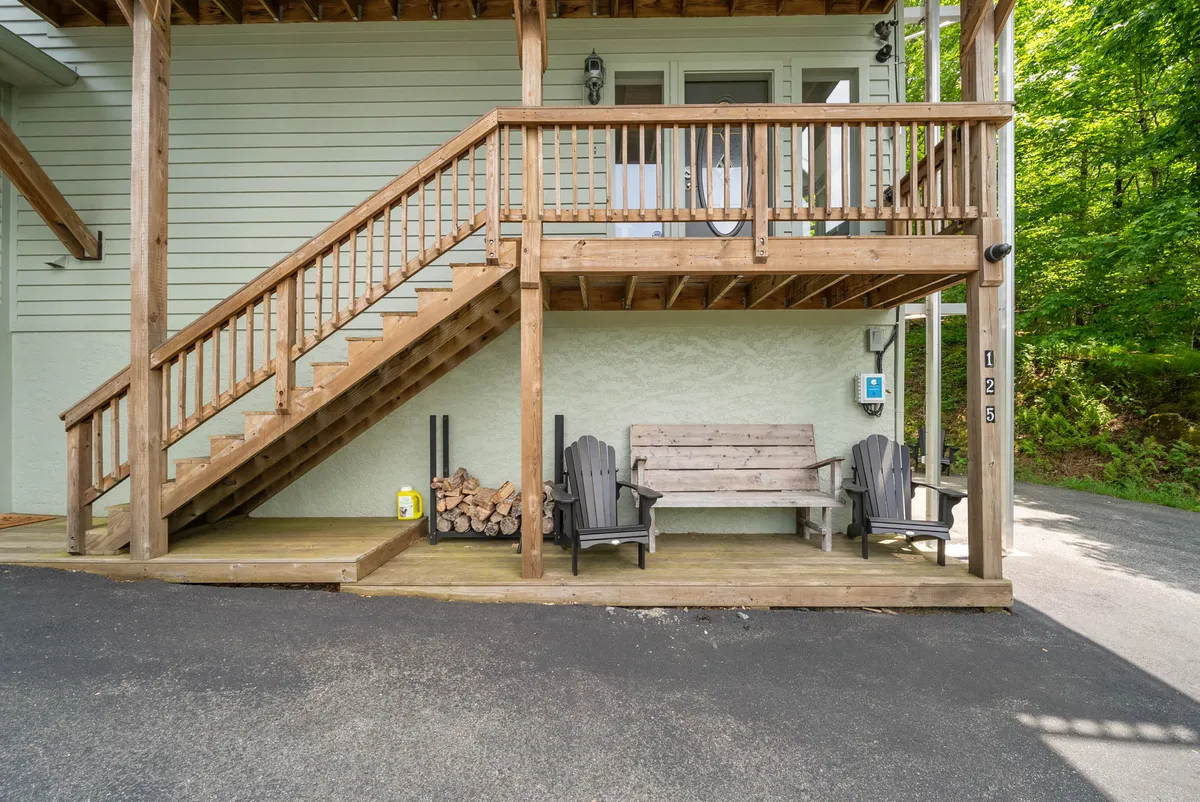 This exterior shot features a welcoming wooden staircase leading up to a cozy seating area surrounded by greenery. A wood pile adds to the rustic charm, and the deck looks perfect for casual gatherings or relaxing after a day of adventure.