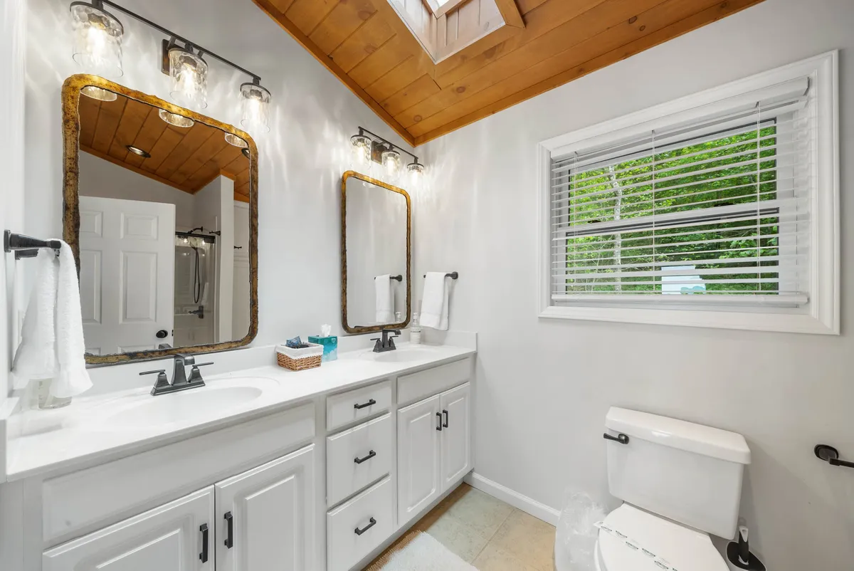 A modern, spa-inspired bathroom featuring a double vanity with mirrors, a skylight that brings in natural sunlight, wooden ceilings, and elegant fixtures. The space feels airy, clean, and perfect for self-care routines.