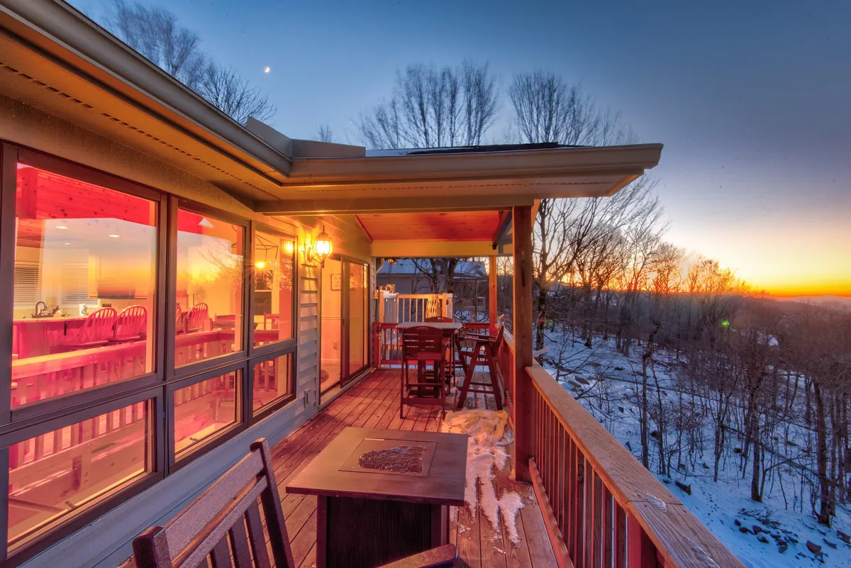 A cozy deck seating area is shown at sunset, with warm interior light glowing through the windows. A fire pit is set up for evening gatherings, offering an inviting transition between indoor comfort and outdoor enjoyment.