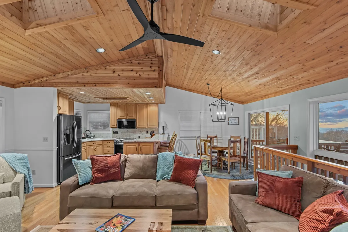 A wide view of an open-concept living space showing the living area with a comfortable sofa, the dining area with a wooden table and chairs, and a fully visible kitchen space in the background. Warm wooden ceilings and large windows flood the room with natural light, and the furniture is arranged to encourage gathering and togetherness.