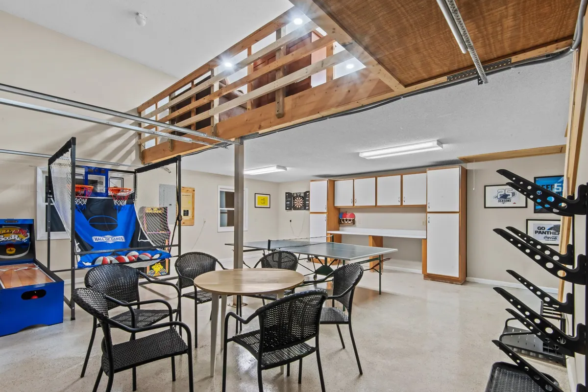 This image showcases the vibrant garage game room, featuring a basketball arcade game, a ping pong table, and a comfortable seating area. The upper loft is visible in the background, highlighting the multi-level fun and social space. The scene captures the energetic, playful mood of the space, with clear views of family-friendly amenities.