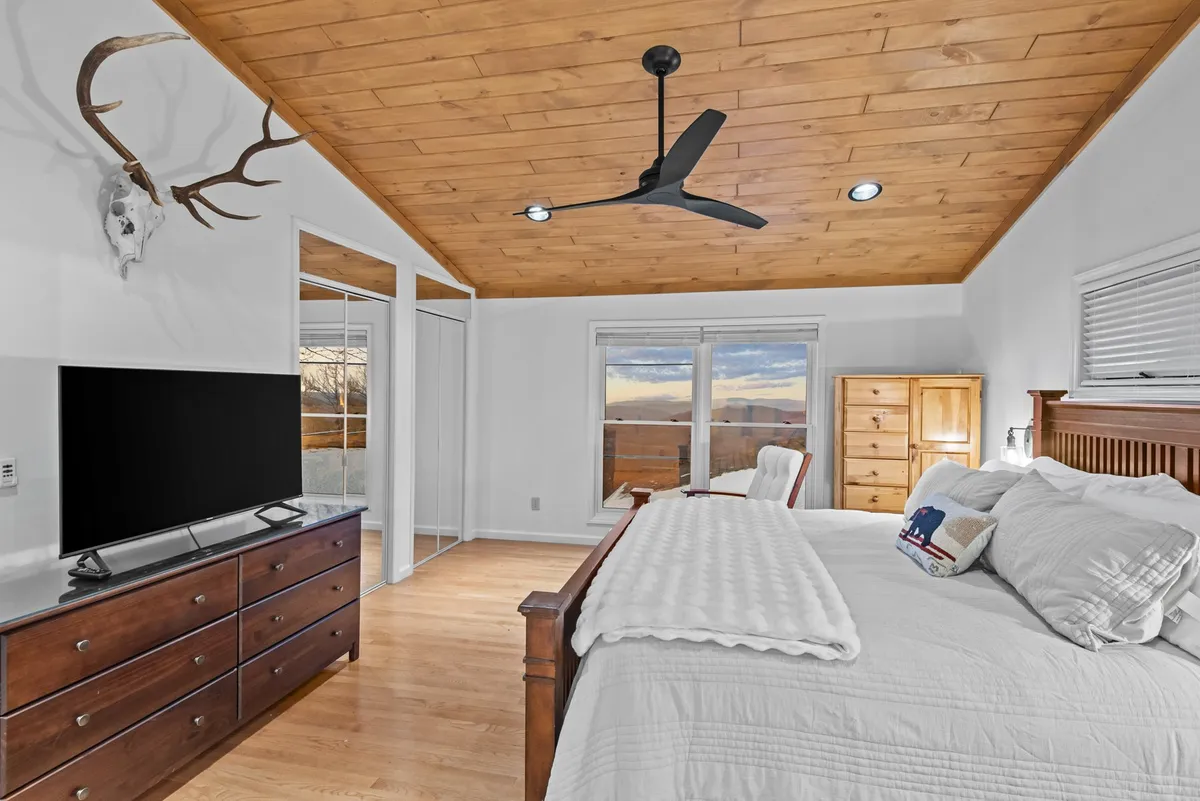A tranquil bedroom with a soft, white-linen bed, a wooden dresser, a TV unit, and antler-themed decor. Natural light filters through, creating a restful ambiance perfect for a peaceful night's sleep or a quiet morning in.