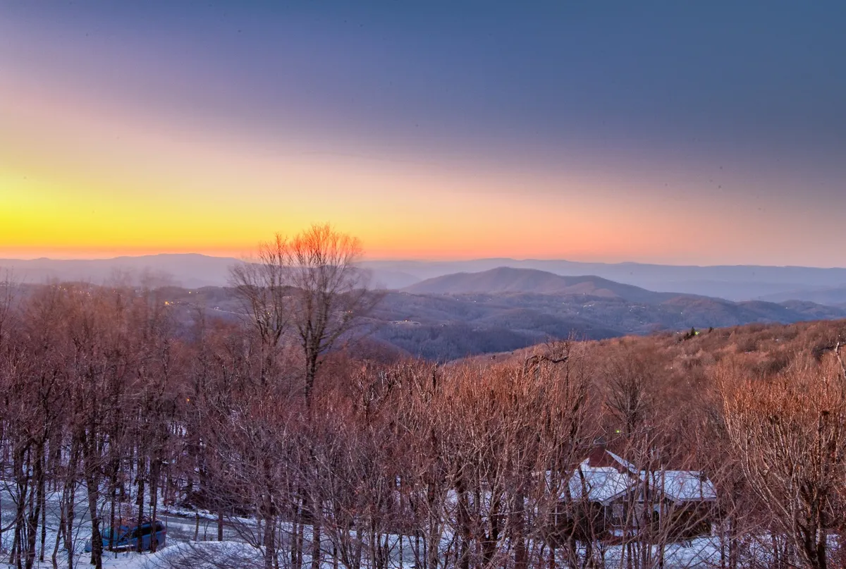 A breathtaking view of a mountain range at sunset, with bare trees framing the vista and a sky painted in rich twilight colors. The landscape suggests serenity and opportunity for activities like hiking, photography, and stargazing.