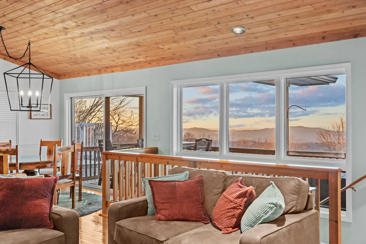 A welcoming dining and living area featuring a wooden dining table with chairs, plush seating with decorative pillows, and large windows offering a beautiful scenic view. The space is bathed in natural light, highlighting the inviting, communal atmosphere.
